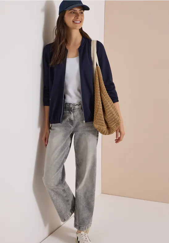 Femme en tenue décontractée avec veste bleu marine, t-shirt blanc, jean ample gris et sac tressé à l’épaule.