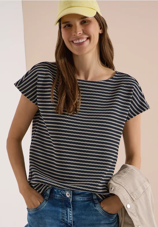 Vrouw in gestreept T-shirt en spijkerbroek, houdt een lichte jas vast voor een neutrale achtergrond.