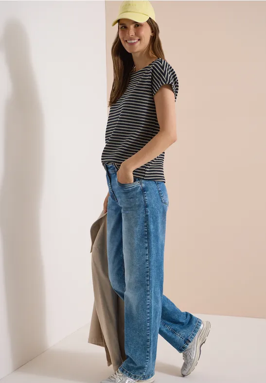 Een vrouw in een gestreept t-shirt, wijde jeans en sneakers staat nonchalant met een jas in haar hand.