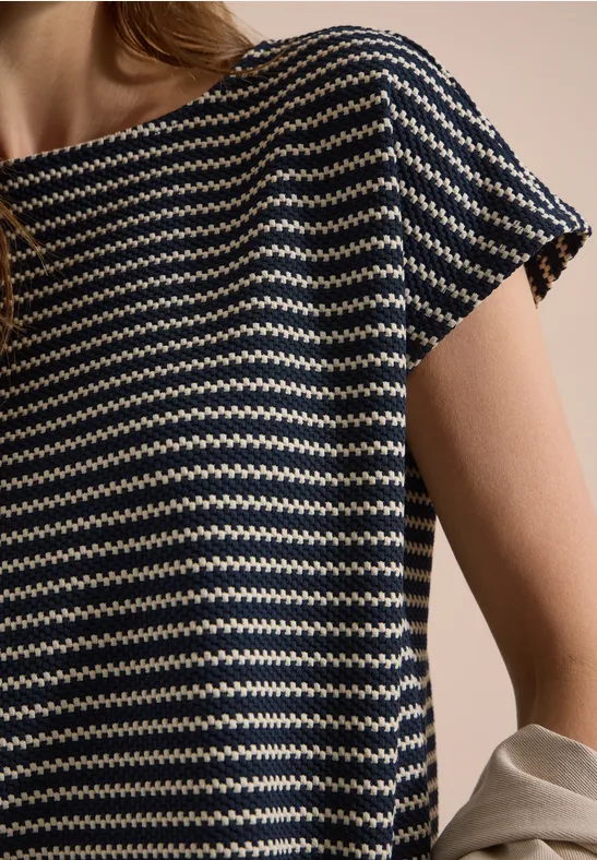 Persoon draagt een marineblauw shirt met witte zigzagstrepen en korte mouwen, met een beige jasje voor neutrale achtergrond.