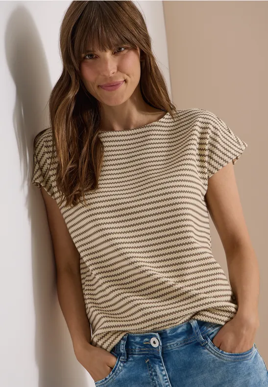 Een vrouw leunt tegen een muur en draagt een beige T-shirt met strepen en een blauwe spijkerbroek.
