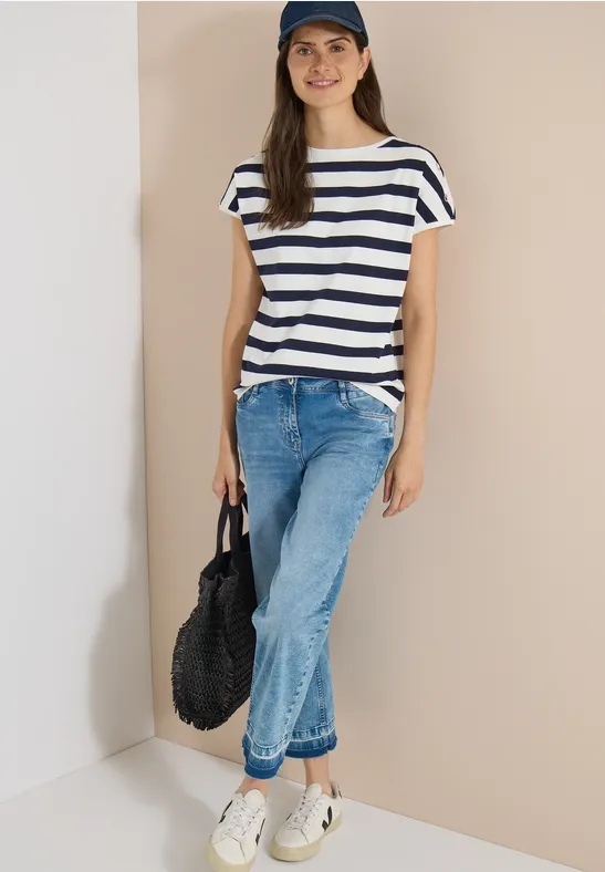 Personne en t-shirt rayé bleu-blanc, jean, baskets blanches et casquette tenant un sac noir devant un fond clair.
