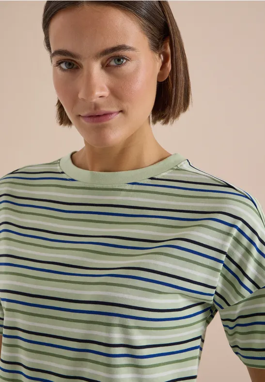 Een vrouw met kort bruin haar draagt een groen-blauw gestreept T-shirt voor een beige achtergrond.