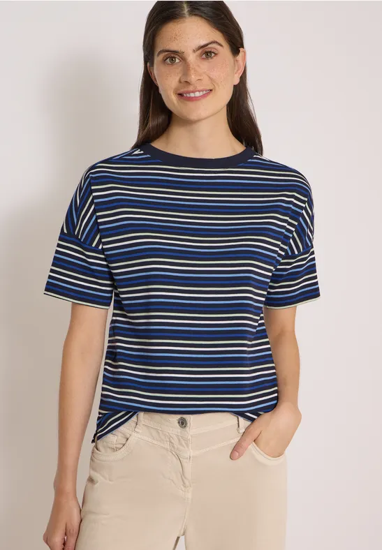 Vrouw glimlacht in blauw-wit gestreept T-shirt en beige broek, met één hand in de zak.