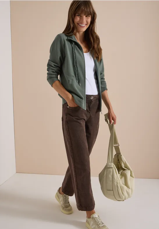 Jongedame lacht, draagt witte blouse, groen jasje, bruine broek, beige tas en beige sneakers.
