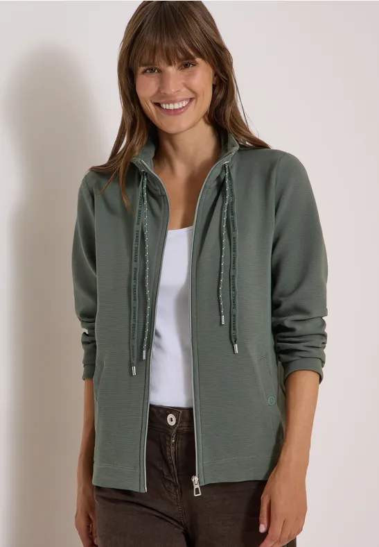 Lachende vrouw met een groene capuchontrui en witte top voor een lichte achtergrond.