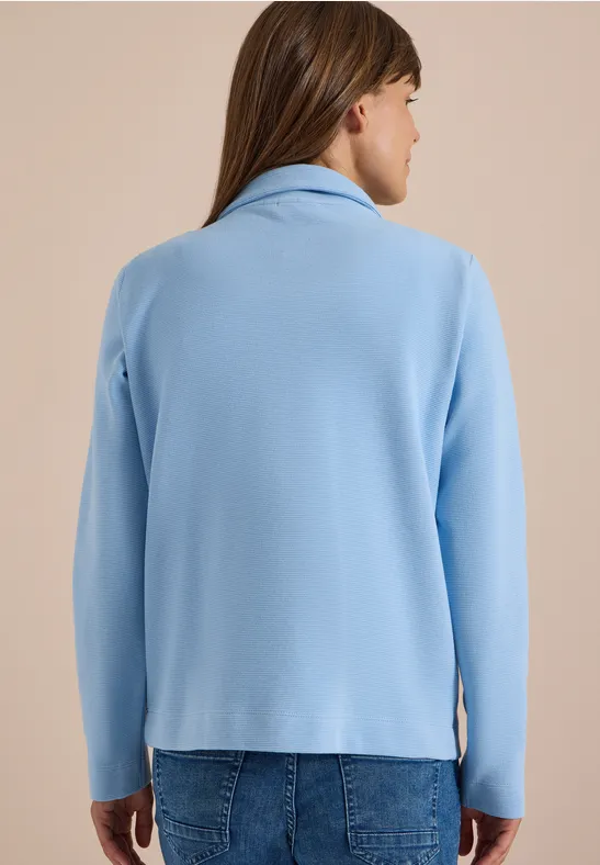 Frau von hinten in hellblauer Jacke und blauer Jeans vor neutralem Hintergrund.