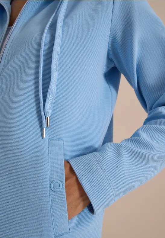 Hand in Seitentasche einer hellblauen Kapuzenjacke mit Reißverschluss und Kordeln.