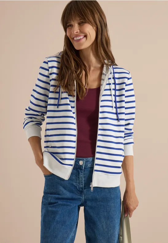 Vrouw glimlachend in witte hoodie met blauwe strepen, bordeaux t-shirt en jeans