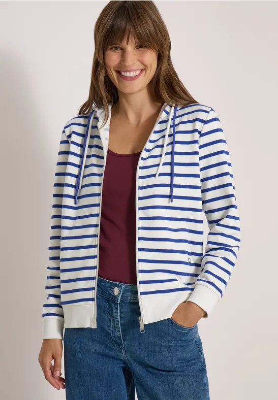 Vrolijke vrouw in een wit met blauwe strepen hoodie, bordeaux top en spijkerbroek.