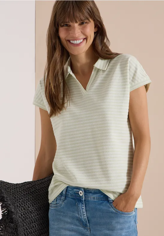 Une femme souriante porte un polo rayé vert pâle à col V et un jean.