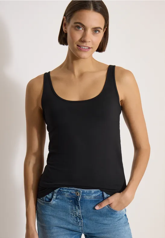 Femme en débardeur noir et jean bleu, une main dans la poche, fond neutre.
