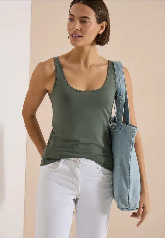 Frau trägt grünes Tanktop, weiße Hose und blaue Stofftasche, vor hellem Hintergrund.