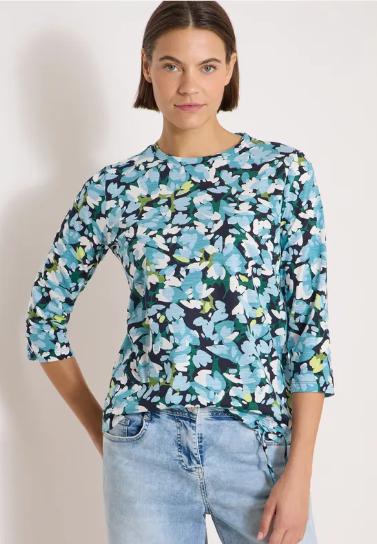 Frau trägt blau-schwarzes Blumen-Shirt mit 3/4-Ärmeln und helle Jeans.