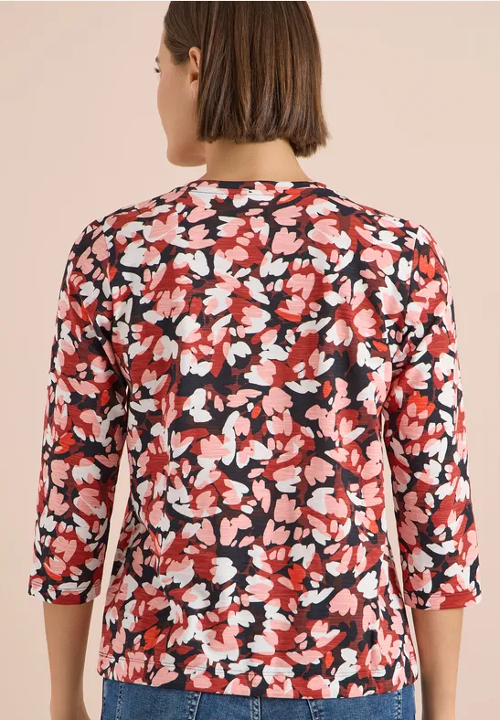 Rückansicht einer Frau in rotem Blusenshirt mit weiß-schwarzem Blumenmuster und 3/4-Ärmeln.