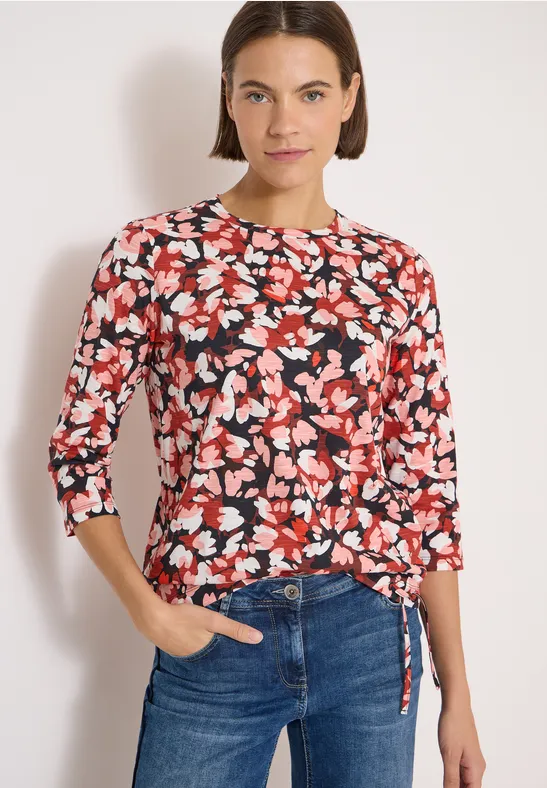 Frau trägt roten, schwarz-weiß Blumenpullover mit 3/4-Ärmeln und Bindeband, kombiniert mit blauen Jeans.
