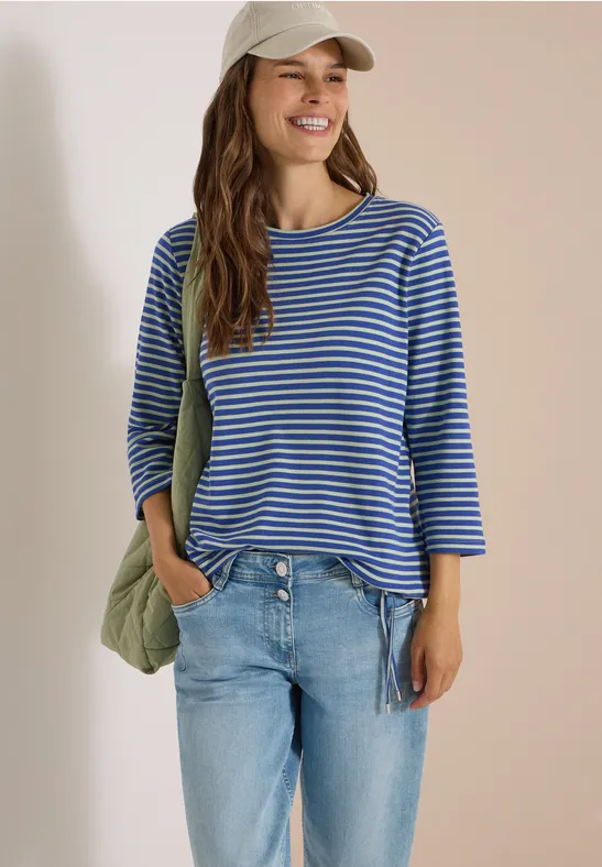 Jeune femme souriante en t-shirt à rayures bleues, jean, casquette et sac en bandoulière.