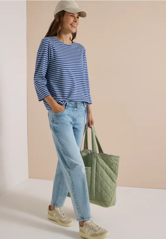 Femme en marinière bleue, jean et baskets blanches, casquette et grand sac matelassé vert.