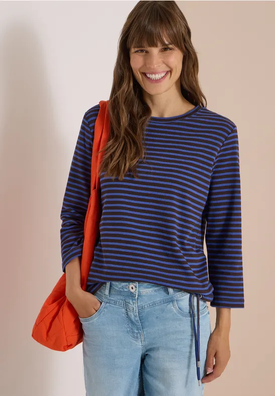 Femme souriante en t-shirt à rayures bleu et noir, jean et sac rouge sur fond clair