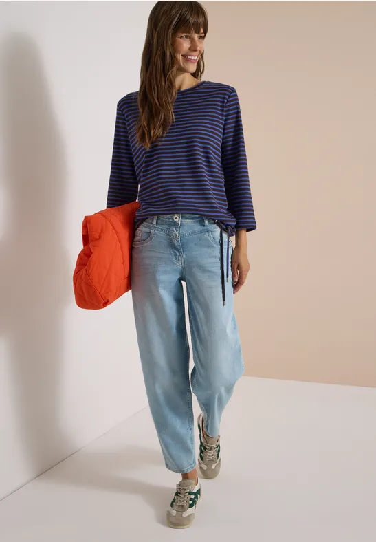 Femme en marinière, jean large et baskets, tient une doudoune orange dans un décor épuré.