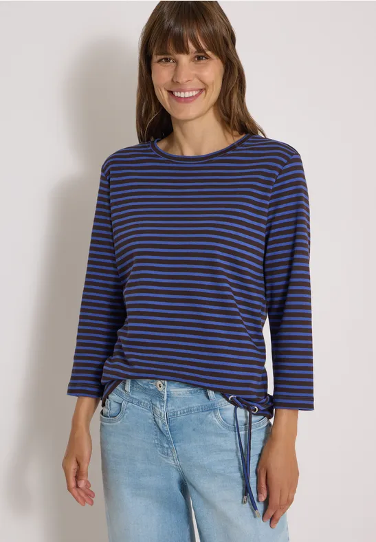 Femme souriante en haut à rayures bleues et jean devant un fond blanc.