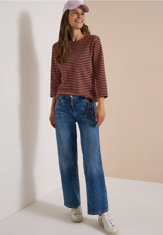 Femme en jean, haut rayé et casquette devant un mur neutre.