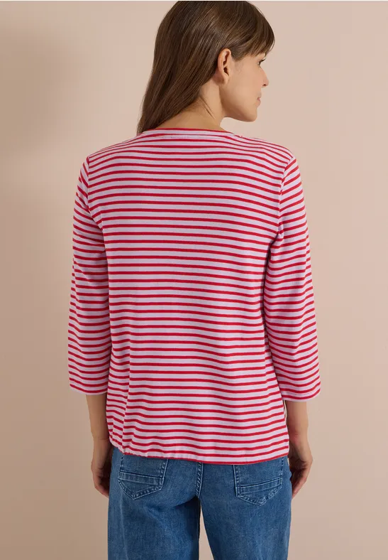 Frau von hinten in Jeans vor hellem Hintergrund, trägt ein pink-weiß gestreiftes Shirt mit 3/4-Ärmeln.