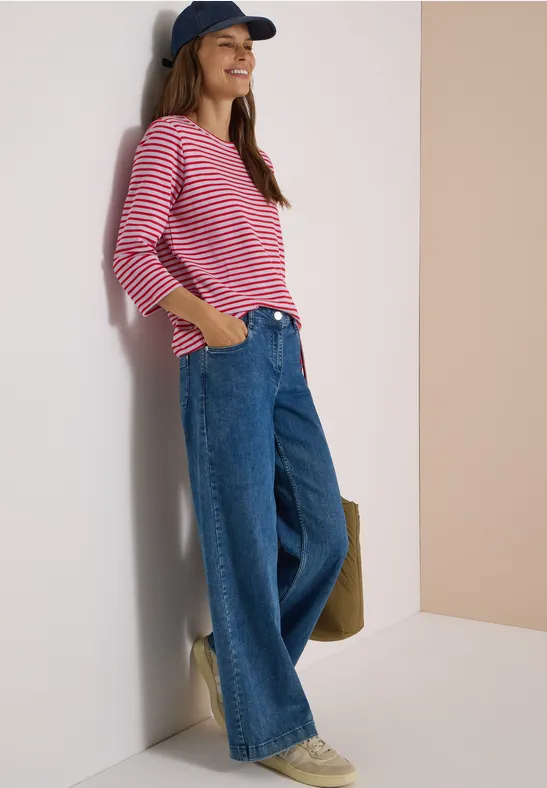Junge Frau lehnt lächelnd an der Wand, trägt gestreiftes Shirt, weite Jeans und Cap.