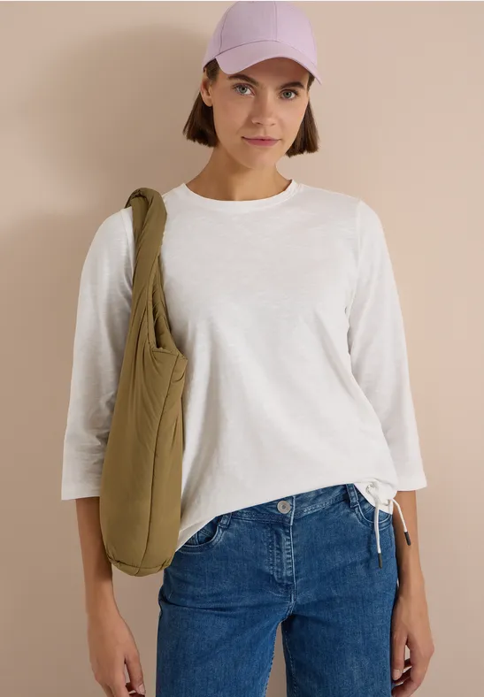 Jeune femme en tee-shirt blanc à manches longues, jeans bleus, casquette violette et sac vert sur fond beige.