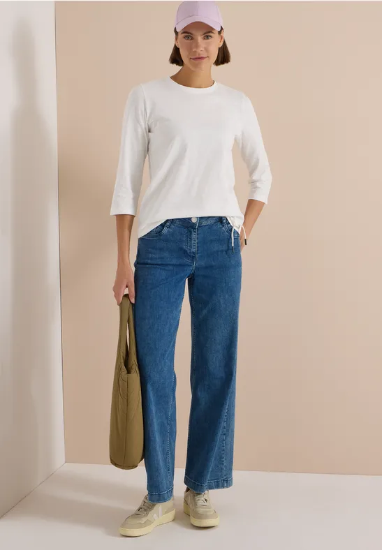 Femme en tee-shirt blanc 3/4, jean bleu, baskets beiges et sac olive, devant un mur beige