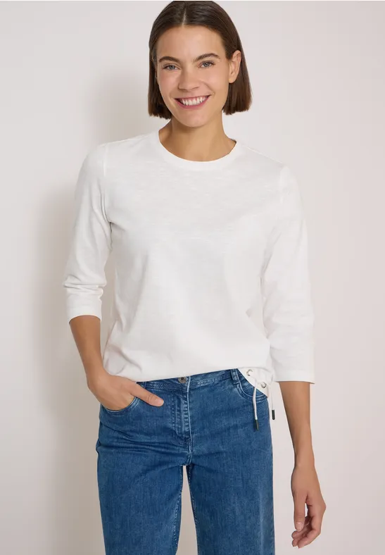 Jeune femme souriante portant un t-shirt blanc à manches longues et un jean bleu devant un fond neutre