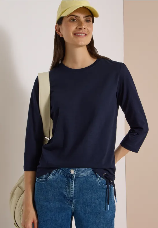 Une femme souriante porte un haut bleu marine à manches 3/4, un jean, une casquette jaune et un sac.
