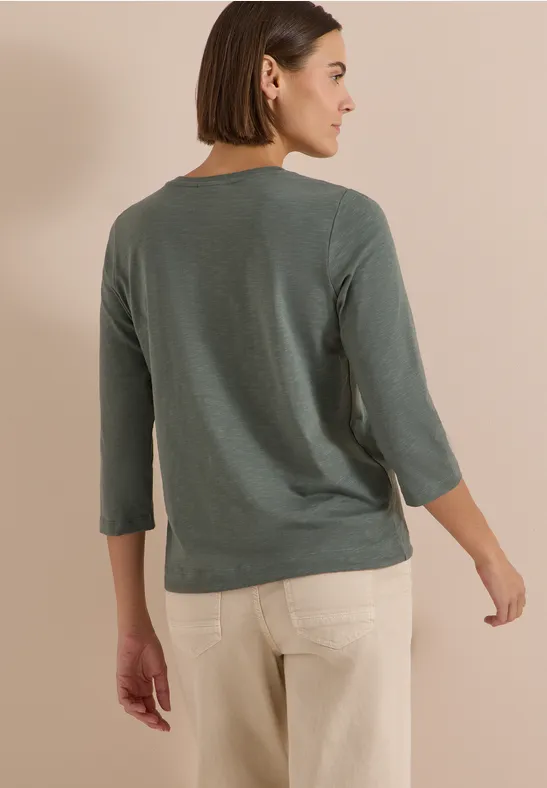 Femme de dos en pull vert 3/4 et pantalon beige devant un mur crème