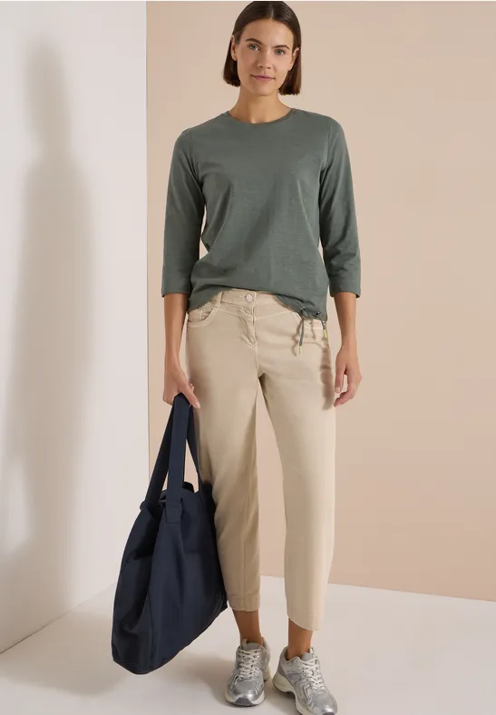 Femme en haut gris à manches trois-quarts, pantalon beige et baskets, tenant un grand sac bleu marine.