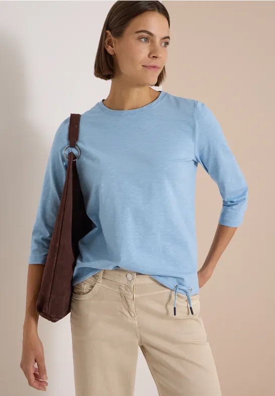 Femme portant un t-shirt bleu clair à manches 3/4, pantalon beige et sac marron, regard latéral sur fond neutre.