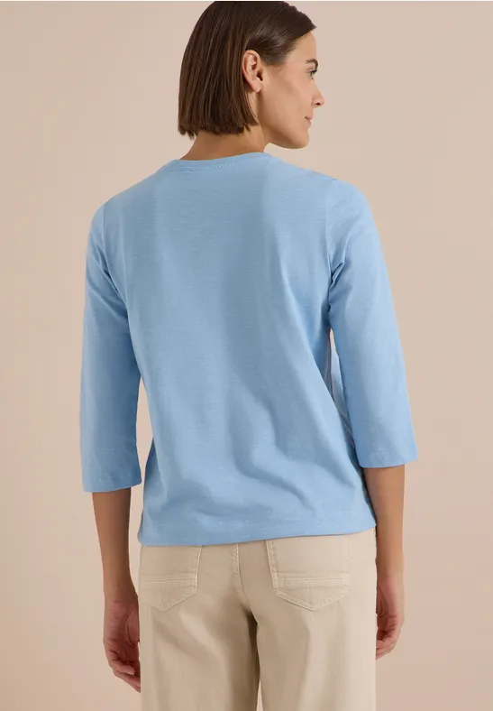 Vue de dos d'une femme portant un haut bleu clair à manches trois-quarts et un pantalon beige.