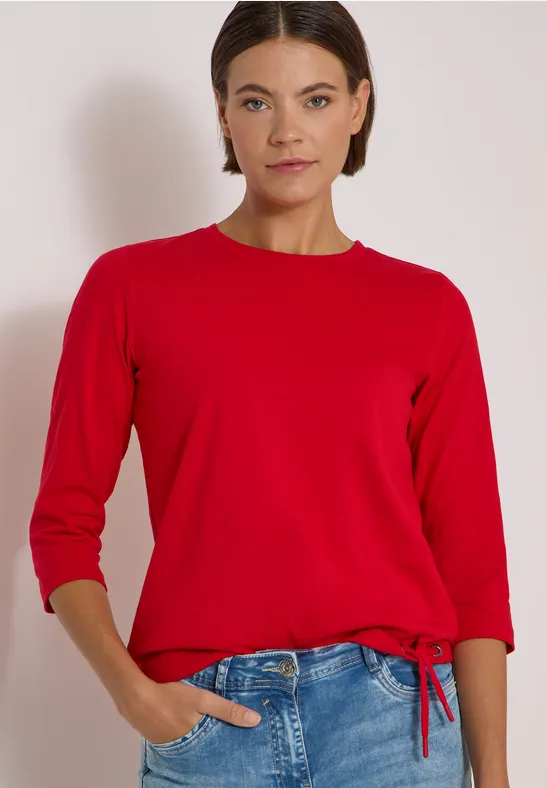 Frau vor hellem Hintergrund trägt roten Pullover mit 3/4-Ärmeln und blaue Jeans, eine Hand in Tasche.