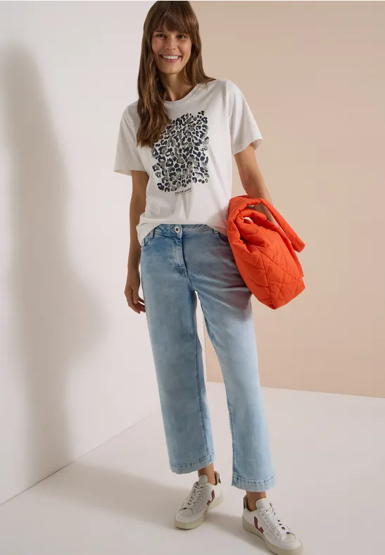 Junge Frau lehnt an Wand, trägt weißes T-Shirt mit Print, helle Jeans, weiße Sneaker und orange Jacke.