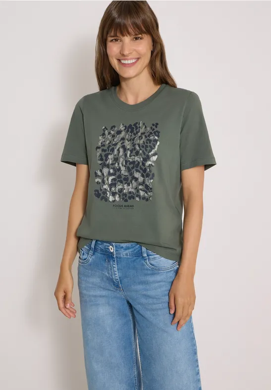 Frau in olivgrünem T-Shirt mit floralem Print und blauen Jeans, lächelnd vor weißem Hintergrund.