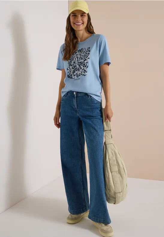 Frau mit blauem T-Shirt und weitem Jeans, gelber Cap und großer Stepp-Tasche.