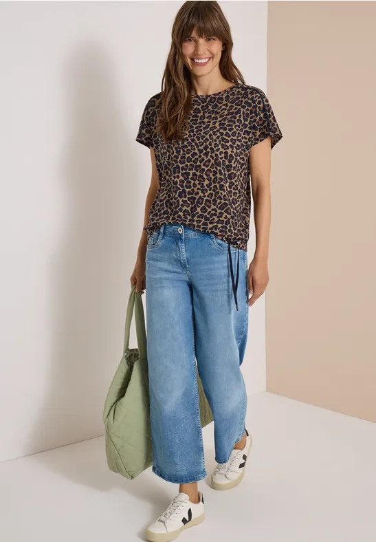Frau trägt Leoparden-T-Shirt, weite Jeans, weiße Sneaker und hält eine grüne Tasche vor hellem Hintergrund.