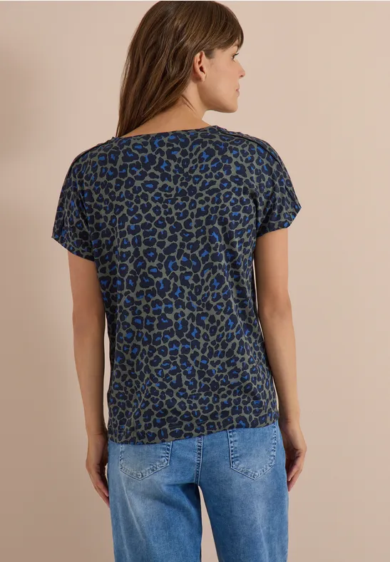Rückansicht einer Frau in blau-schwarz gemustertem T-Shirt und Jeans vor neutralem Hintergrund.