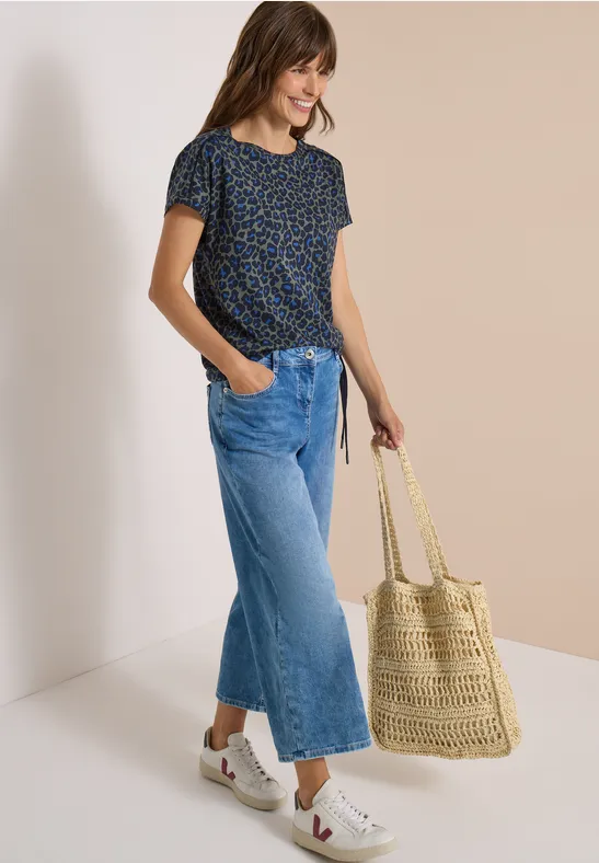 Frau in Leopardenmuster-Shirt, weiten Jeans und Basttasche lächelnd