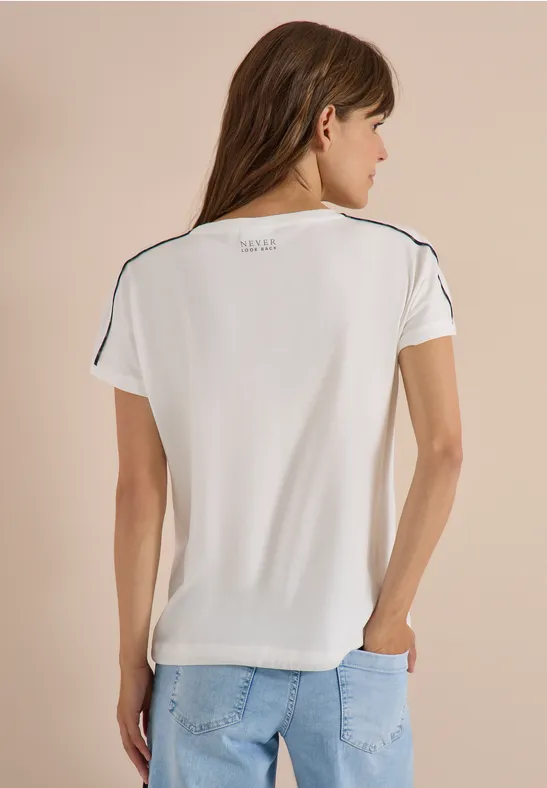 Frau von hinten in weißem Kurzarmshirt mit schwarzen Nähten und Jeans vor neutralem Hintergrund.