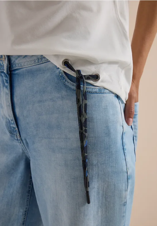 Person trägt hellblaue Jeans mit Camouflage-Kordel, Hand in Tasche und weißem T-Shirt.