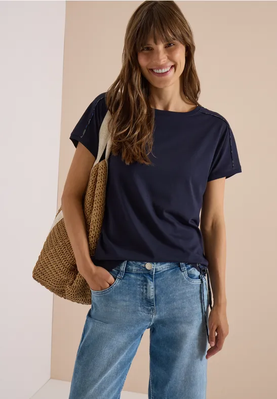 Junge Frau in casual Outfit: dunkelblaues T-Shirt, Jeans und Basttasche, lächelnd vor neutralem Hintergrund.