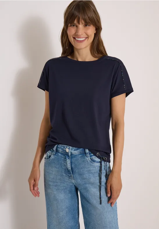 Frau im dunkelblauen T-Shirt mit seitlicher Raffung und blauen Jeans, lächelnd vor hellem Hintergrund.