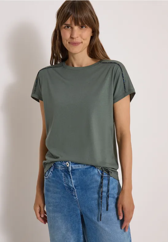 Frau mit braunen Haaren in grünem Kurzarmshirt mit Kordeln und blauen Jeans.