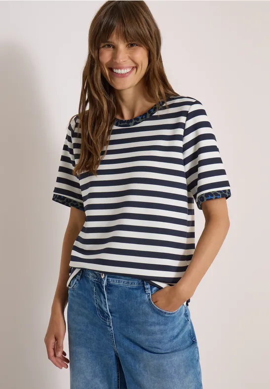 Frau mit braunen Haaren lächelnd, gestreiftes T-Shirt und Jeans vor hellem Hintergrund.