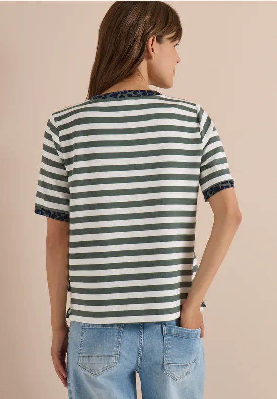Eine Frau von hinten trägt ein grün-weiß gestreiftes Kurzarmshirt mit blauer Spitzenborte an Ärmeln und Saum und Jeans.